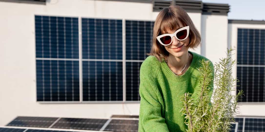 Vrouw op dakterras, zet een pot met plant neer en heeft zonnepanelen op de achtergrond